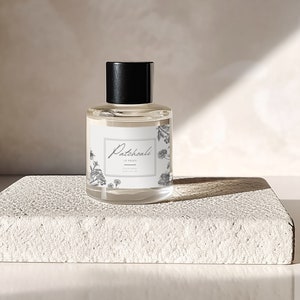 Może przedstawiać: Przezroczysta butelka szklana z czarną zakrętką i białą etykietą z napisem "Patchouli Le Parfum". Butelka stoi na białej, teksturowanej powierzchni.