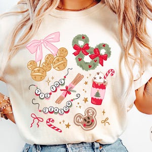 以下が含まれることがあります： アイボリー色のTシャツ。クリスマスをテーマにしたお祝いのグラフィックが特徴です。ピンクのリボン、ミッキーマウス型のベル、リース、キャンディケイン、ジンジャーブレッドクッキーなどのデザイン。テキスト「TIS THE SEASON」も表示されています。