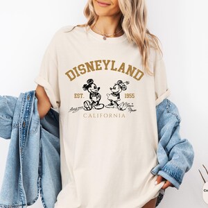 May include: A white t-shirt featuring a brown graphic of Mickey Mouse and Minnie Mouse. The graphic reads "Disneyland Est. 1955 California".