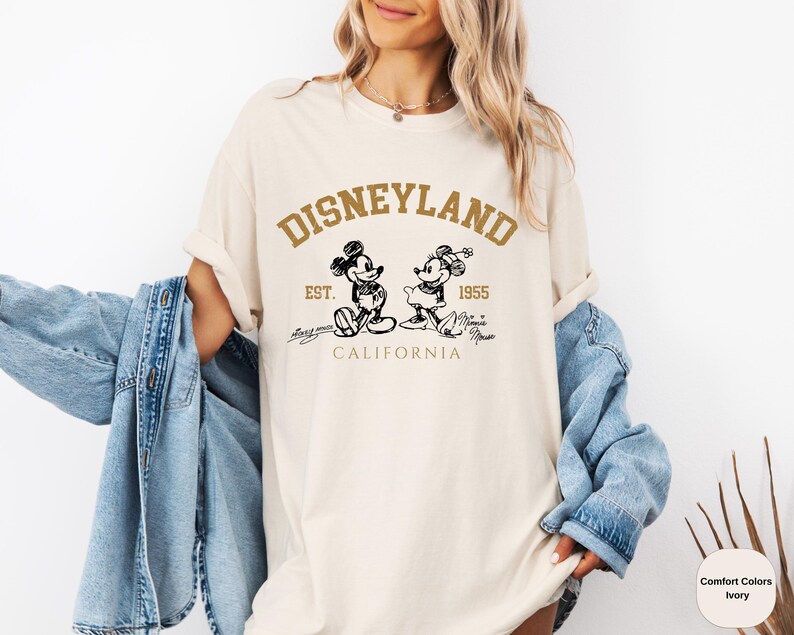 May include: A white t-shirt featuring a brown graphic of Mickey Mouse and Minnie Mouse. The graphic reads "Disneyland Est. 1955 California".
