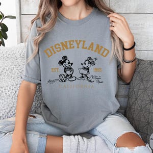 May include: A grey t-shirt with a vintage-style graphic of Mickey and Minnie Mouse in front of the text "DISNEYLAND EST. 1955 CALIFORNIA".