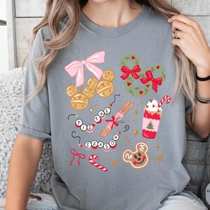 以下が含まれることがあります： お祝いのグラフィックが施されたグレーのTシャツ。デザインには、ピンクのリボン、金のベル、ミッキーマウス型のリース、シナモンスティック、キャンディケーン、ジンジャーブレッドクッキーが含まれています。「Tis the Season」というテキストも表示されています。