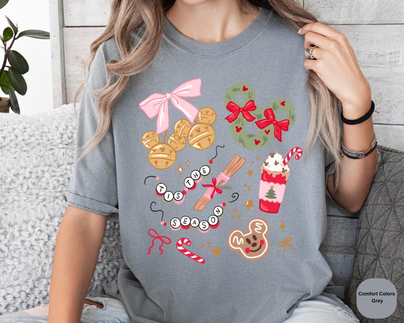 以下が含まれることがあります： お祝いのグラフィックが施されたグレーのTシャツ。デザインには、ピンクのリボン、金のベル、ミッキーマウス型のリース、シナモンスティック、キャンディケーン、ジンジャーブレッドクッキーが含まれています。「Tis the Season」というテキストも表示されています。