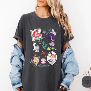 Puede incluir: Una camiseta gris oscuro con un estampado gr&aacute;fico de varios personajes de la pel&iacute;cula de Disney La Sirenita. El gr&aacute;fico incluye a Ariel, &Uacute;rsula, el pr&iacute;ncipe Eric, Sebasti&aacute;n, Flounder y otros personajes. El texto de la camiseta dice "Mermaid at Heart", "Sea Ya Later", "Salty But Sweet" y "Make a Splash".