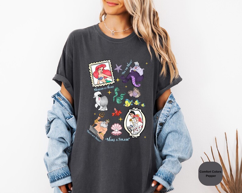 Puede incluir: Una camiseta gris oscuro con un estampado gr&aacute;fico de varios personajes de la pel&iacute;cula de Disney La Sirenita. El gr&aacute;fico incluye a Ariel, &Uacute;rsula, el pr&iacute;ncipe Eric, Sebasti&aacute;n, Flounder y otros personajes. El texto de la camiseta dice "Mermaid at Heart", "Sea Ya Later", "Salty But Sweet" y "Make a Splash".