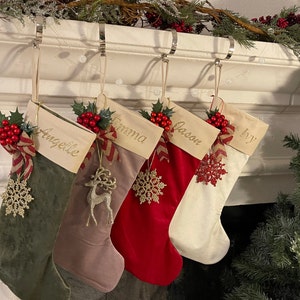 May include: Five Christmas stockings hang from a mantel. Each stocking is a different color: green, brown, red, cream, and white. Each stocking has a name embroidered on it, along with decorative accents like snowflakes and reindeer.