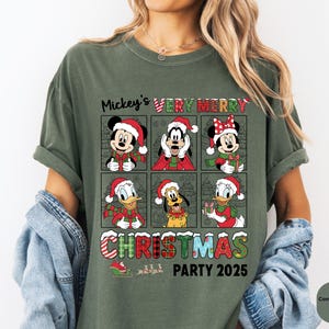 Puede incluir: Camiseta verde oliva con un dise&ntilde;o festivo. Presenta a Mickey Mouse, Minnie Mouse, Goofy, Donald Duck y Pluto, todos con gorros y bufandas de Pap&aacute; Noel. El texto dice "Mickey's Very Merry Christmas Party 2025".