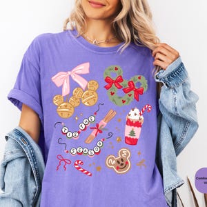 以下が含まれることがあります： 紫色のComfort Colors Tシャツ。お祝いのホリデーグラフィックが特徴です。デザインには、ミッキーマウス型のベル、リース、ジンジャーブレッドクッキー、ホリデー ドリンクが含まれています。「'Tis the Season」というフレーズも表示されています。