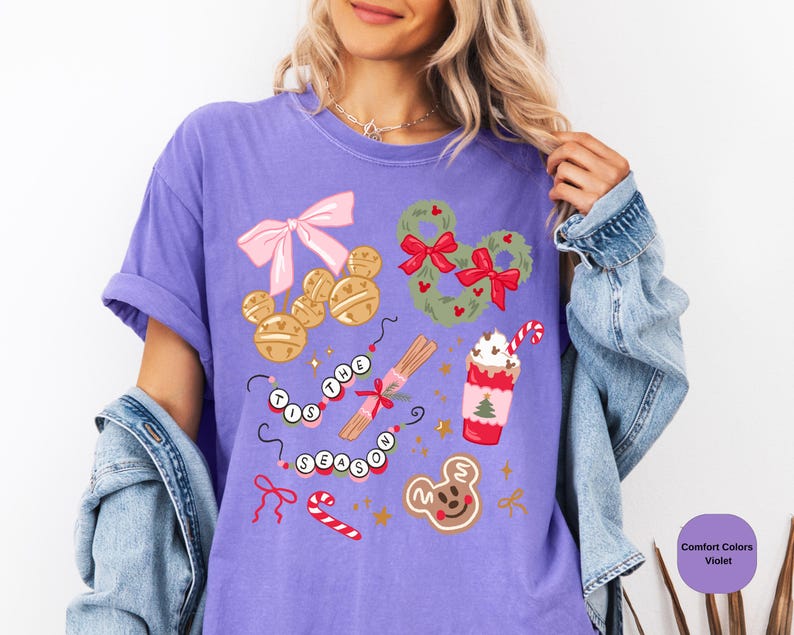 以下が含まれることがあります： 紫色のComfort Colors Tシャツ。お祝いのホリデーグラフィックが特徴です。デザインには、ミッキーマウス型のベル、リース、ジンジャーブレッドクッキー、ホリデー ドリンクが含まれています。「'Tis the Season」というフレーズも表示されています。