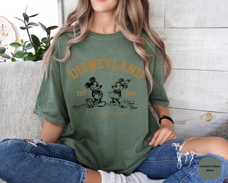 May include: A green t-shirt with the text 'DISNEYLAND EST. 1955 CALIFORNIA' in gold lettering. The t-shirt features a black and white illustration of Mickey Mouse and Minnie Mouse.