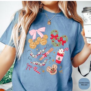 以下が含まれることがあります： 青いジーンズカラーのTシャツ。お祝いのグラフィックデザイン。ミッキーマウスの形をしたオーナメント、リース、ジンジャーブレッドクッキー、ホイップクリームのカップが含まれています。「TIS THE SEASON」の文字もデザインされています。