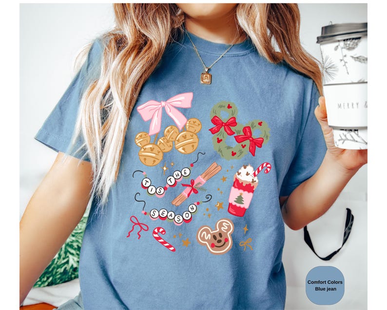 以下が含まれることがあります： 青いジーンズカラーのTシャツ。お祝いのグラフィックデザイン。ミッキーマウスの形をしたオーナメント、リース、ジンジャーブレッドクッキー、ホイップクリームのカップが含まれています。「TIS THE SEASON」の文字もデザインされています。