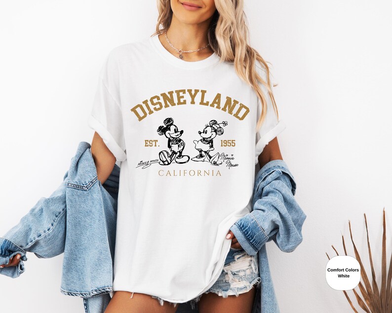 May include: White t-shirt with a gold graphic of Mickey Mouse and Minnie Mouse in front of the text "DISNEYLAND EST. 1955 CALIFORNIA".