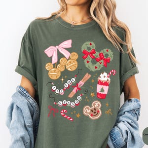 以下が含まれることがあります： モスグリーンのTシャツ。ピンクのリボン、ミッキーマウスのベルオーナメント、リース、キャンディケーン、ホットチョコレートなどのお祝いのグラフィックが特徴です。「'Tis the Season」と書かれています。