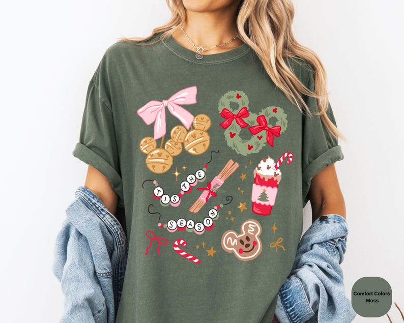 以下が含まれることがあります： モスグリーンのTシャツ。ピンクのリボン、ミッキーマウスのベルオーナメント、リース、キャンディケーン、ホットチョコレートなどのお祝いのグラフィックが特徴です。「'Tis the Season」と書かれています。