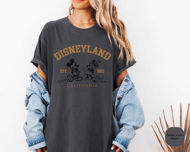 May include: A dark grey t-shirt with a vintage-style graphic of Mickey Mouse and Minnie Mouse in front of the Disneyland logo. The text reads "DISNEYLAND EST. 1955 CALIFORNIA".