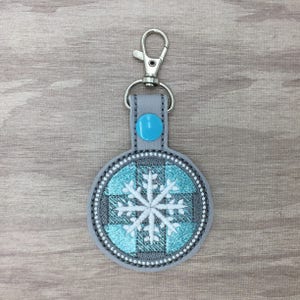 May include: A gray and blue snowflake keychain with a silver clip. The snowflake is embroidered on a round, gray fabric base with a blue and white plaid pattern.