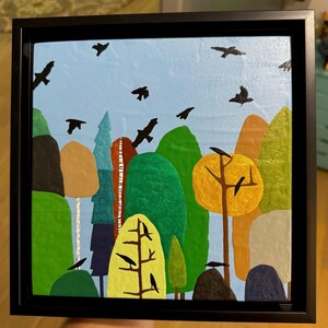 May include: A framed painting featuring a whimsical landscape with trees in shades of green, brown, and yellow against a light blue sky. Black birds are scattered throughout the scene. The artwork is enclosed in a black frame.