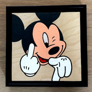 May include: A framed artwork featuring a cartoon mouse giving the middle finger. The mouse has black ears, a peach-colored face, and a red mouth. The artwork is set against a light wood grain background and framed in black.