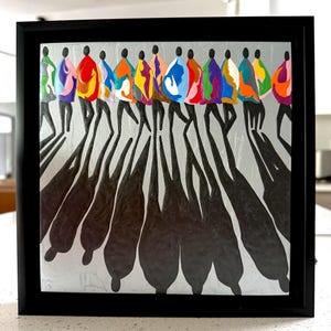 May include: Abstract art print in a black frame. The artwork features a line of silhouetted figures in colorful robes, casting long black shadows on a silver background. The figures are depicted with simple shapes and bold colors.