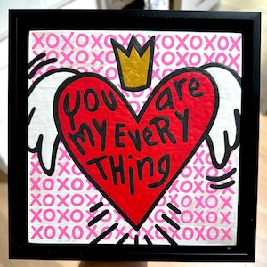 May include: Framed artwork with a red heart at the center, inscribed with the words "You are my every thing" in black. The heart is crowned with a gold crown. The background is pink with repeating "XOXO" patterns. The artwork is in a black frame.