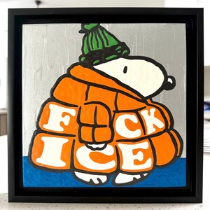 May include: Framed artwork featuring a cartoon character in an orange jacket and green hat. The jacket has the words "FUCK ICE" in white block letters. The background is silver with a blue base, all within a black frame.