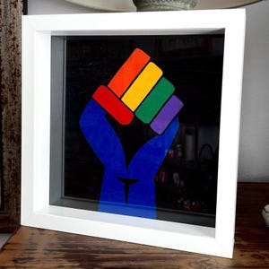 May include: A framed artwork featuring a raised fist in the colors of the rainbow flag. The fist is composed of red, orange, yellow, green, and purple segments, with a blue arm and a black background. The frame is white.