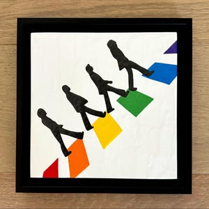 May include: A framed art print featuring four black silhouettes walking on colorful geometric shapes. The figures are set against a white background, with a black frame. The shapes are red, orange, yellow, green, blue, and purple.