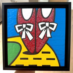 May include: A square framed painting featuring a pair of glittery red shoes with white bows, positioned above a yellow path with black rectangular markings, set against a bright blue background with a green section on the left side of the frame.