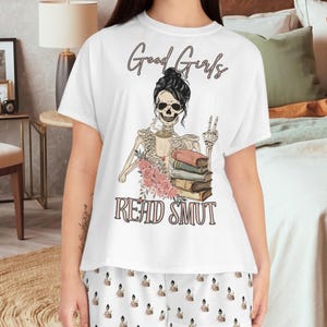 Może przedstawiać: Biały zestaw piżamy z grafiką szkieletu i tekstem "Good Girls Read Smut". Szkielet trzyma stos książek i filiżankę kawy. Spodnie piżamy mają powtarzający się wzór grafiki szkieletu.