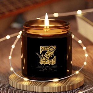 Peut inclure: Un bocal en verre brun contenant une bougie avec une étiquette noire. L'étiquette présente un motif floral doré avec la lettre "Z" au centre. Le texte sur l'étiquette indique "Dare Romance Series, Sable & Rose, Candle Collection".
