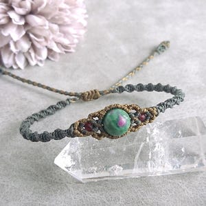 May include: Macrame bracelet with a central green and pink gemstone, and small red beads. The bracelet is made with olive green and tan macrame cord. The bracelet is displayed on a clear crystal with a soft grey background.