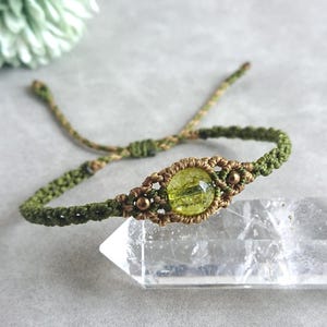 May include: A handmade macrame bracelet with a central, round, light green gemstone. The bracelet is crafted with olive green and tan coloured cord, with a sliding knot closure. The gemstone is approximately 1.3 cm in diameter.