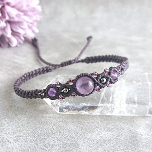 May include: A macrame bracelet crafted from dark grey cord, featuring a central round purple gemstone flanked by two smaller purple stones and metallic beads. The design includes intricate knotting patterns and adjustable cord ends.