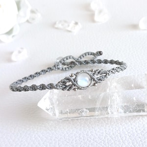 Puede incluir: Una pulsera de macramé gris con una piedra central redonda e iridiscente. La pulsera se muestra sobre un cristal transparente, con detalles blancos en el fondo. El diseño tejido de la pulsera es visible.