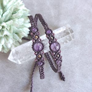 May include: Two macrame bracelets crafted from dark gray cord, each featuring a central round purple amethyst bead, smaller accent amethyst beads, and tiny metallic gold-toned beads woven into the intricate knotted pattern of the cord.