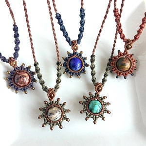 May include: Five macrame necklaces with different coloured cords and gemstone pendants. Each pendant is a sunburst design with a round gemstone in the centre. The gemstones are blue, brown, green, and white.