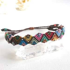 May include: A handmade macrame bracelet featuring a repeating geometric pattern in shades of blue, pink, green, and yellow. The bracelet is secured with a brown cord and is displayed on a clear crystal. This bohemian style bracelet is a unique accessory.