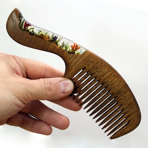 May include: A wooden comb with a handle that has a clear resin section filled with pressed flowers. The comb has a brown wood finish.