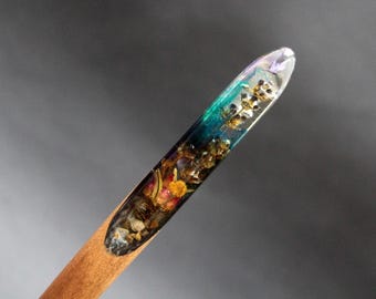 Puede incluir: Un taco de madera con una punta de resina transparente que contiene flores secas y purpurina. La resina es un degradado de turquesa, azul y morado.