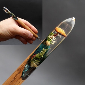 Op de afbeelding: Een houten pen met een transparant harsgedeelte dat een kleine paddenstoel en andere botanische elementen bevat. De pen wordt in een hand gehouden.