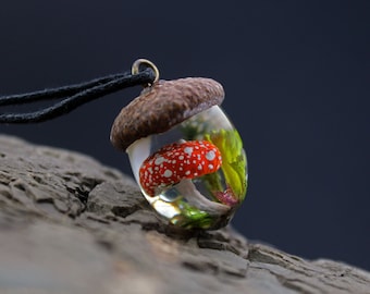 May include: A necklace with a clear acorn pendant containing a red and white mushroom with green foliage.