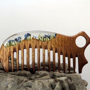 May include: A wooden comb with a clear resin section containing pressed blue flowers. The comb has a handle with a hole in it.