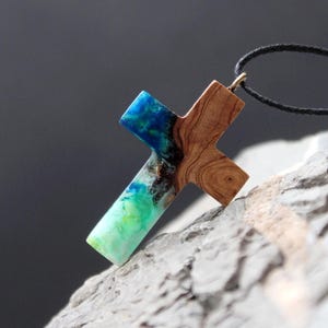 May include: A wooden cross pendant with a black cord. The cross features a combination of wood and resin. The resin portion is a mix of blue, green, and white, while the cross arms are a light brown color.
