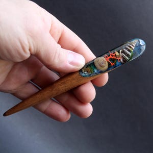 Puede incluir: Un palo de pelo de madera con una sección de resina transparente que contiene conchas marinas y coral. La resina es de color azul claro con toques de verde y naranja.