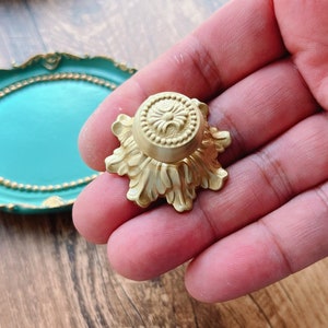 May include: A gold-coloured decorative stand with a round top and intricate floral details. The stand has a raised centre with a circular design.