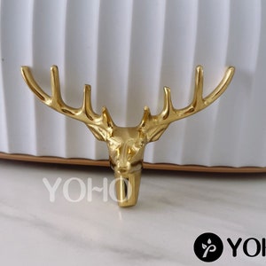 May include: Gold-colored metal deer head ornament with antlers. The deer head is facing forward with its mouth closed. The antlers are curved and point upwards.