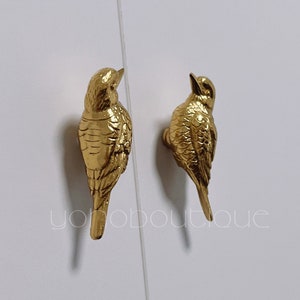 May include: Two gold bird-shaped cabinet knobs. The birds are facing opposite directions. The knobs are detailed with feathers and have a realistic appearance.
