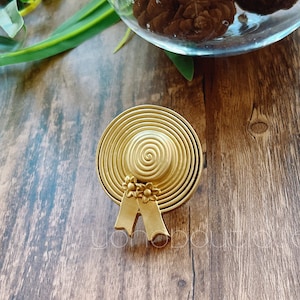 Könnte beinhalten: Eine goldfarbene Metallstift in Form eines Strohhutes mit Schleife und floralen Akzenten.