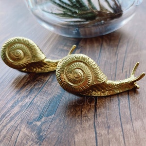 May include: Two gold snail sculptures with detailed shell designs. The snails are on a wooden surface.